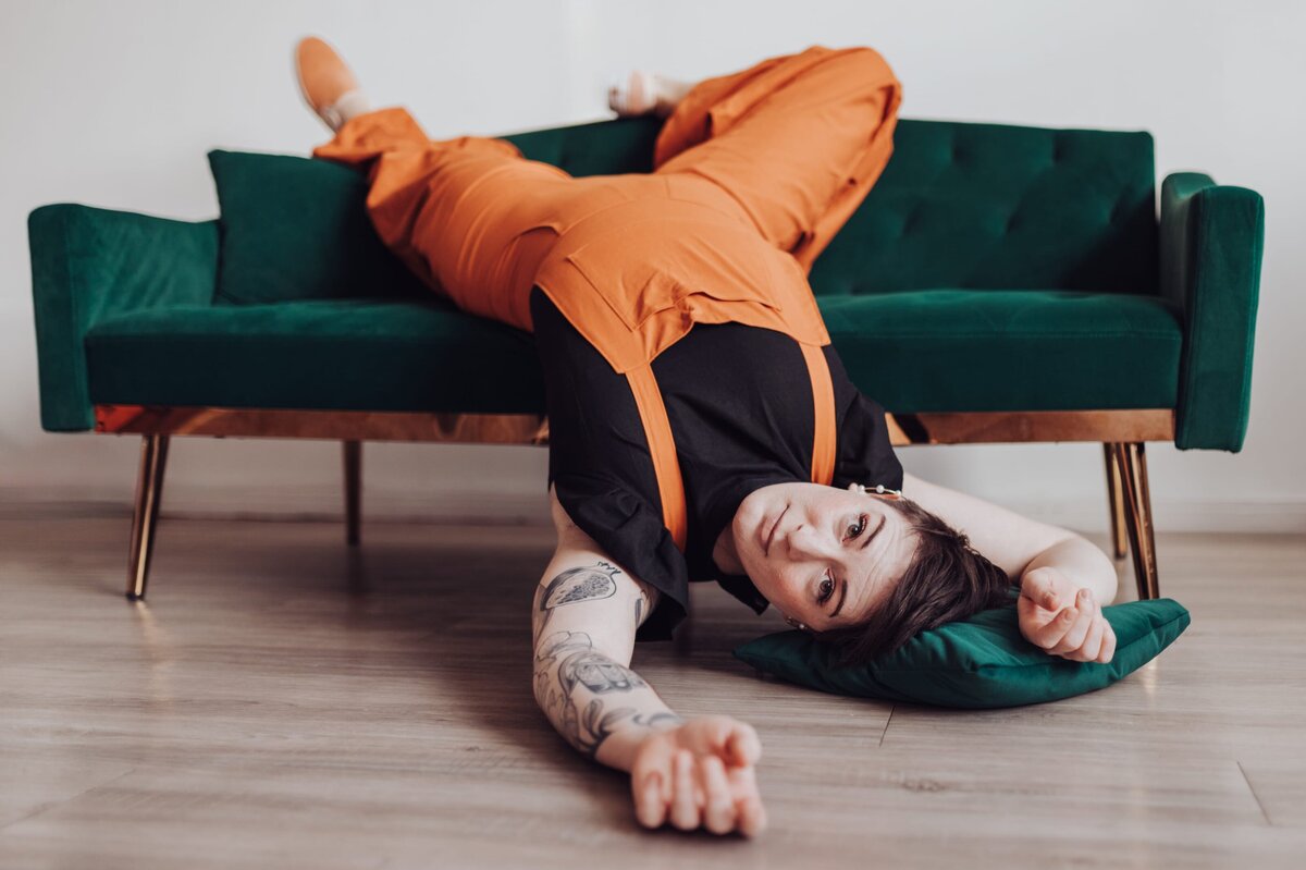 Woman wearing an orange jumpsuit lays upside down wih her head on a green velvet pillow on the floor and her legs over a green velvet sofa as she looks at the camera.