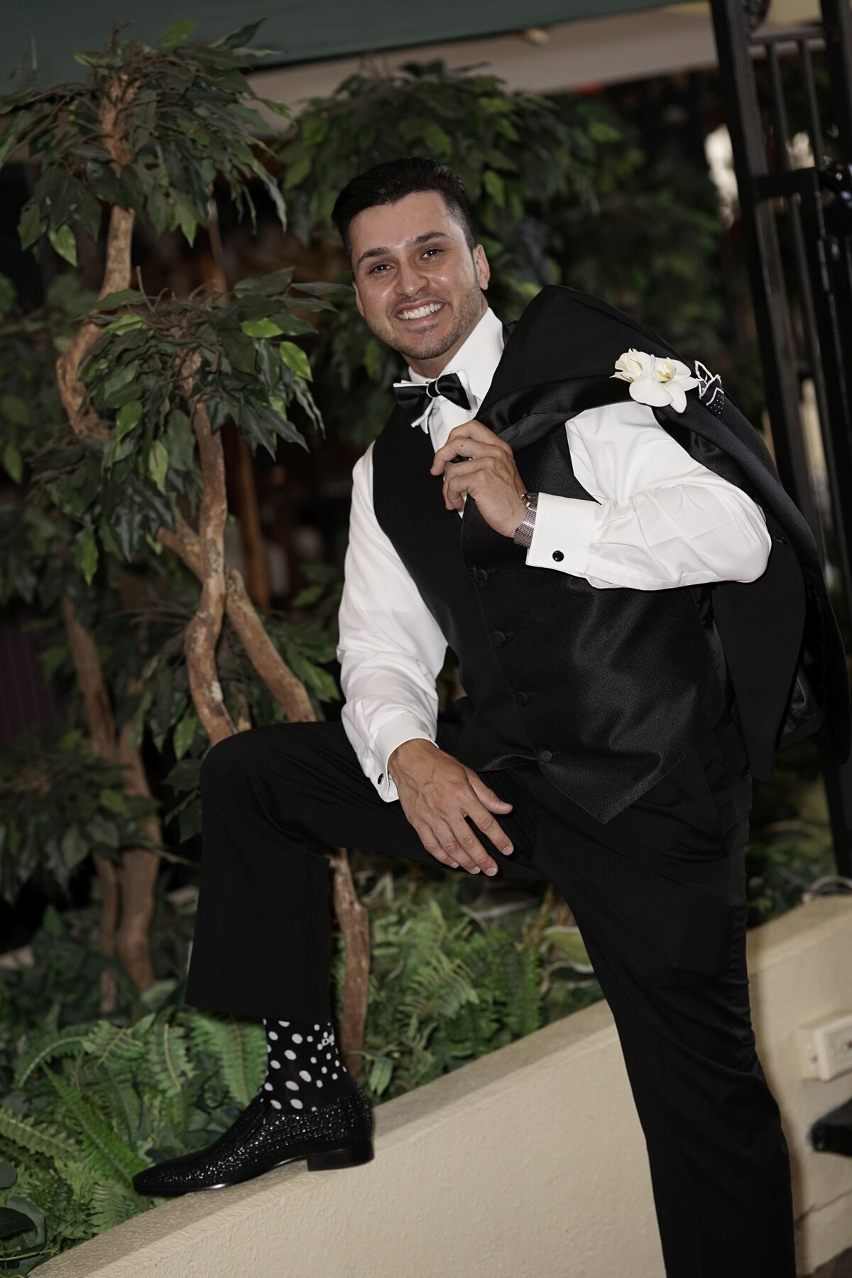 The groom holds his jacket casually over his shoulder while leaning on his knee, showcasing a relaxed yet stylish pose. This image captures a blend of sophistication and ease, emphasizing the groom's confident and elegant demeanor on his wedding day.