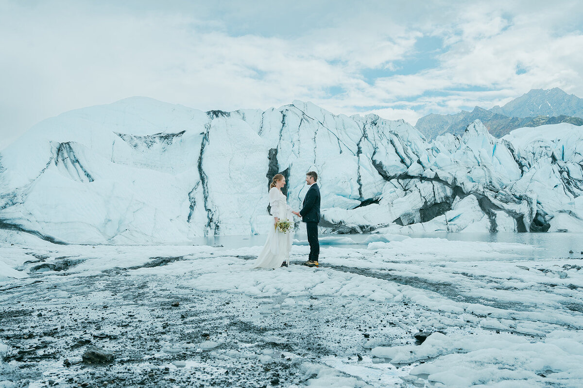 A man and woman in wedding clothing stand face to face holding hands in front of a glacier. 