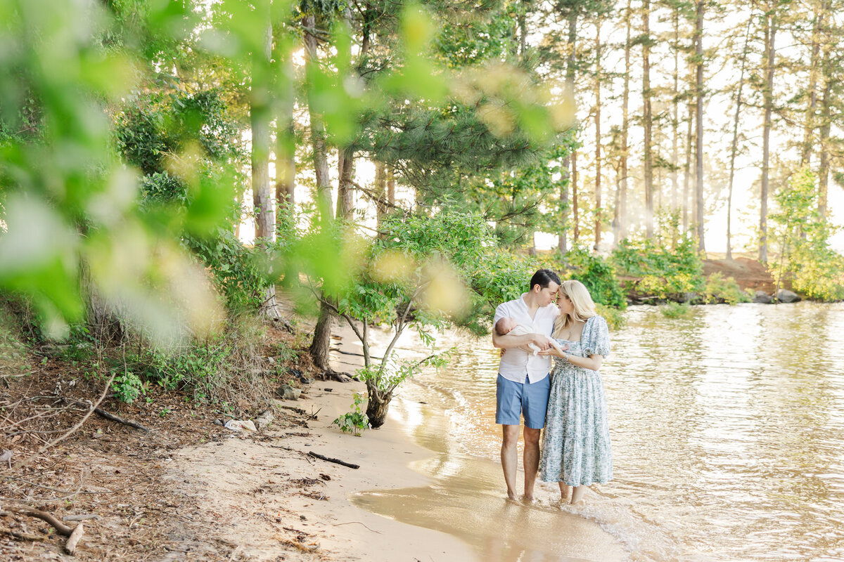 Mom and dad stand in Lake Lanier holding onto newborn baby at sunset.