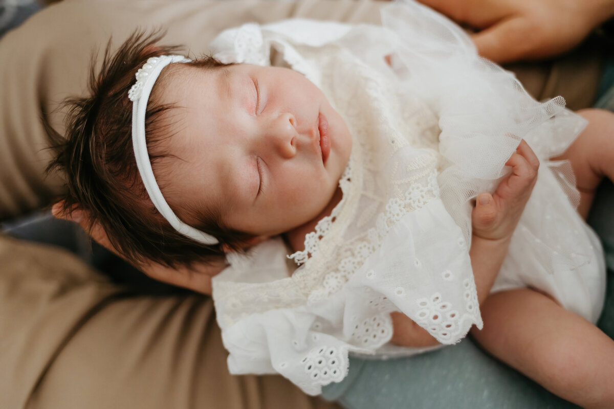 baby girl in white lace outfit and white headband sleeping in her father's arm during a newborn session in costa mesa california