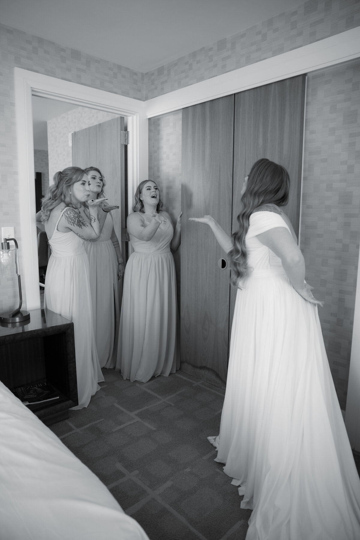 Bride sharing her first look with her bridesmaids, who are smiling and cheering as they see her wedding gown for the first time in an elegant room.