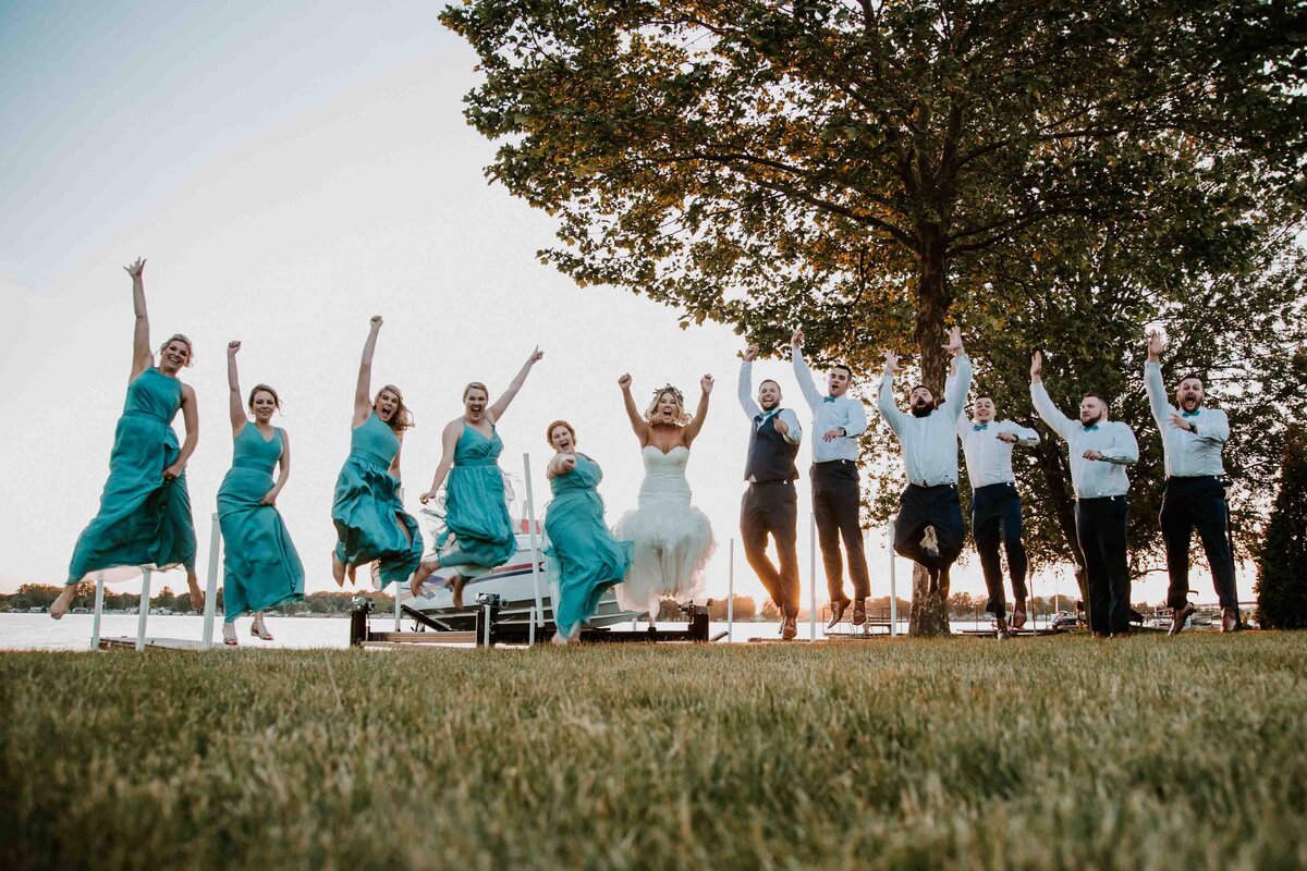 Wedding party jumping in a line on grass in front of Lake Huron. All are excited and happy. Bride and Groom in the middle.