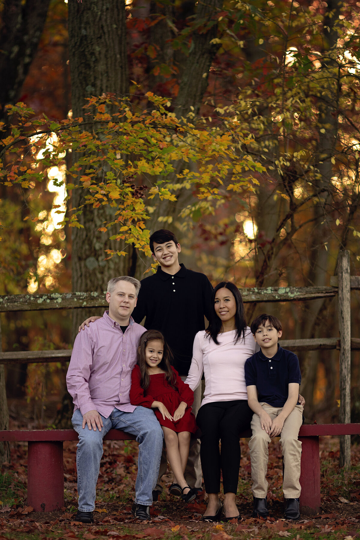 A mother and father sit on a bench with their young son and daughter while their teen stands behind them in a park in fall planned by a New Jersey Family Photographer