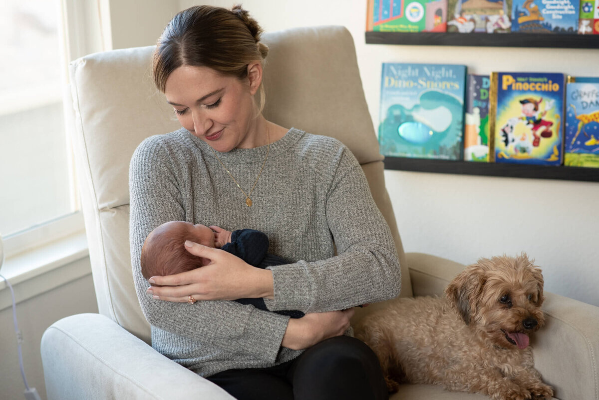 A mother sits on a nursing chair with her small dog while cradling her sleeping newborn