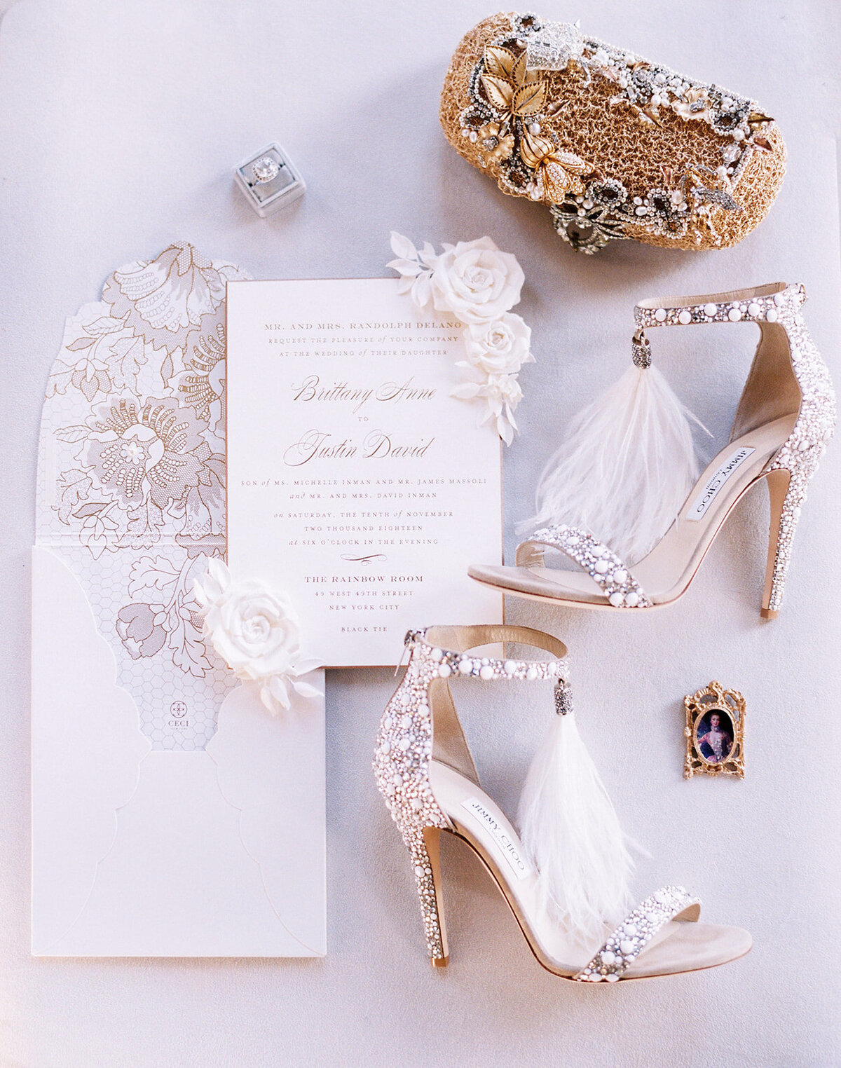 The photo was taken at Paris, France - Chateau Vaux le Vicomte, one of the top destination wedding venues in France, captured by photographer Audrey FORA. The beautifully arranged flat lay display features a wedding invitation suite, a pair of embellished high-heeled shoes with sparkling details and white feathers, an ornate gold clutch intricately designed, and floral decorations. East West Films Videographers were also present at the event to capture stunning footage of the day.