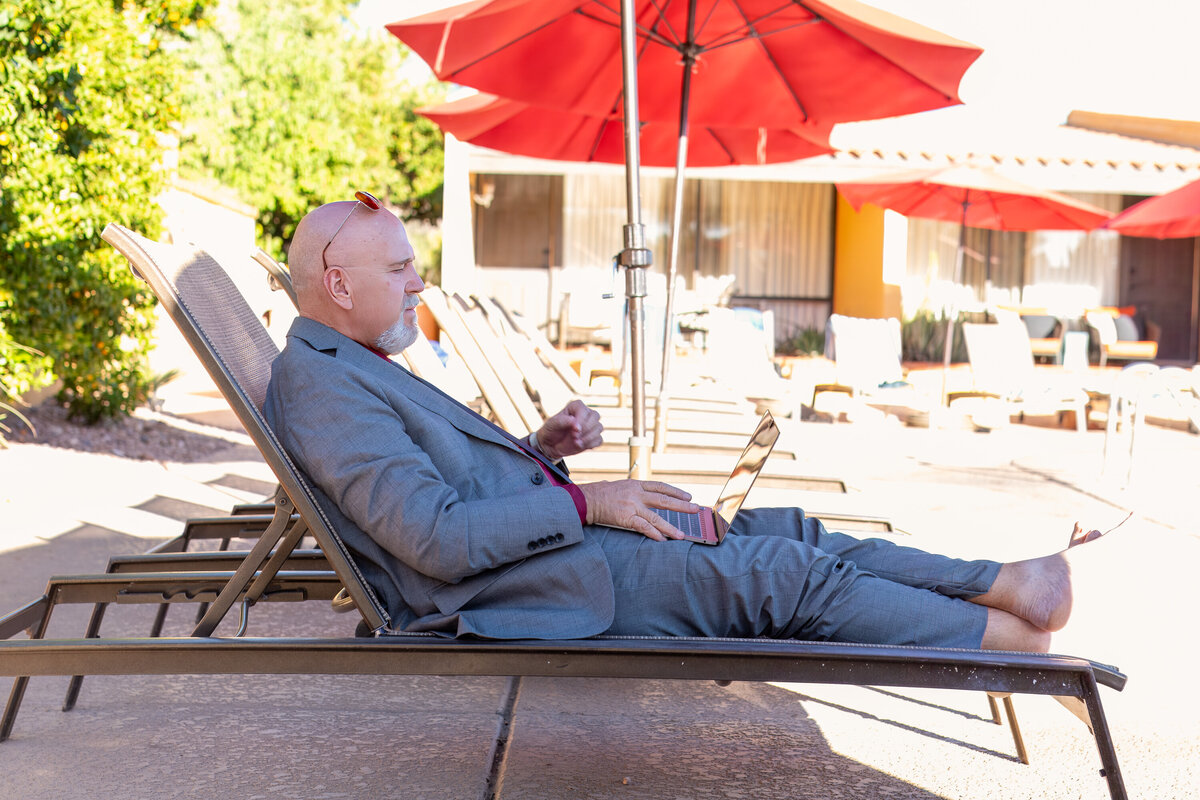 Man in a suit reclining on a poolside lounge chair under red umbrellas while working on a laptop, photographed by Vyrl Photo to showcase a relaxed Tucson business lifestyle scene.