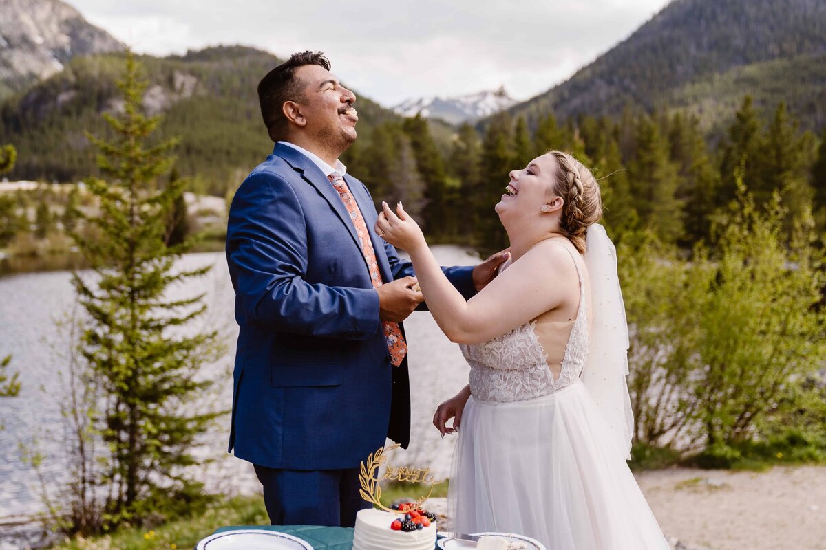 Bride and groom laugh while feeding each other cake during their lakeside Colorado elopement.