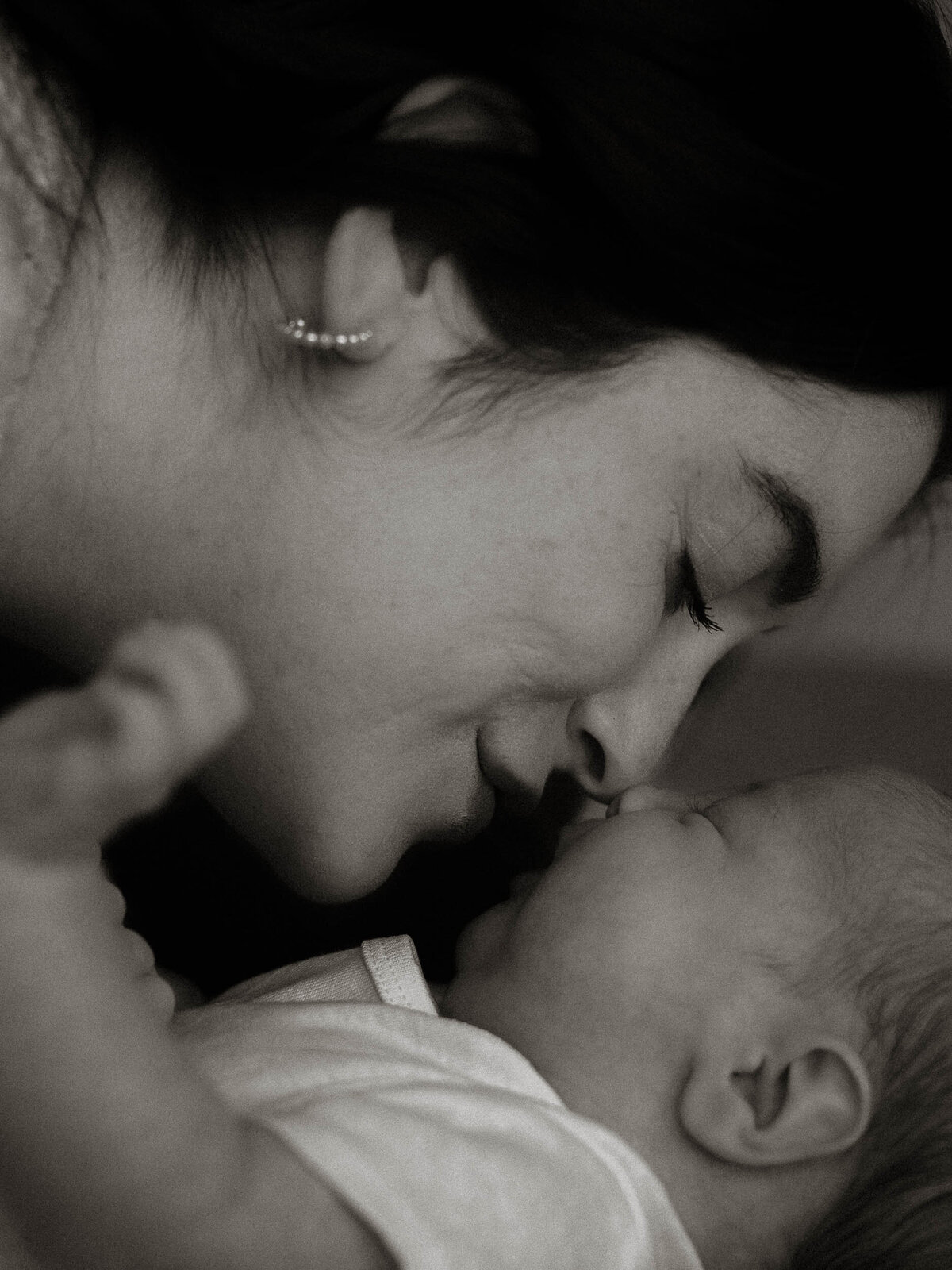 Black and white newborn photography portrait of a mother kissing her baby, intimate and emotional moment during an in-home session.