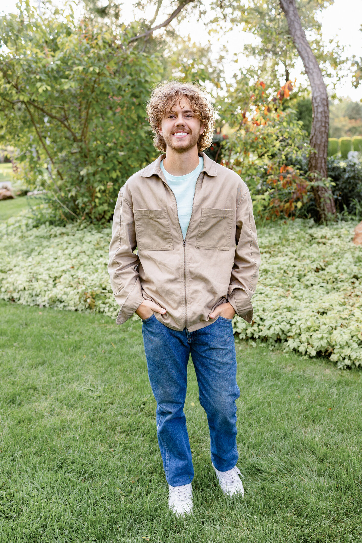 A young man in a beige jacket and jeans stands confidently on a green lawn, smiling at the camera.