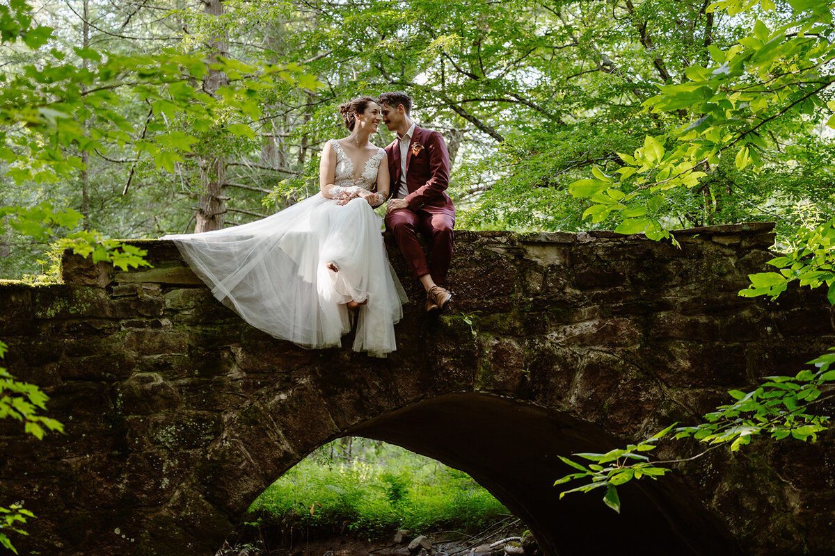 bride and groom sitting on an old stone bridge in the forest