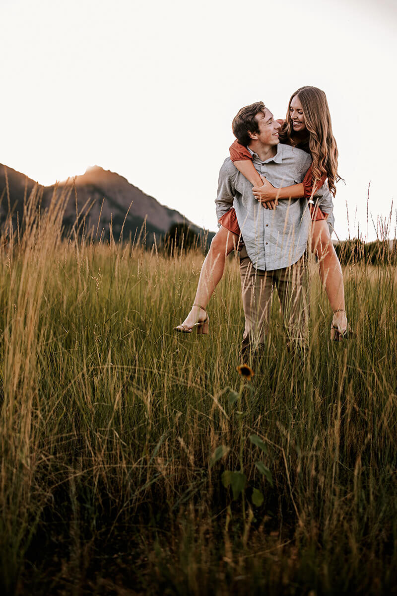 Wife gets piggy back ride from husband in a grassy field with mountains in the background on a summer day