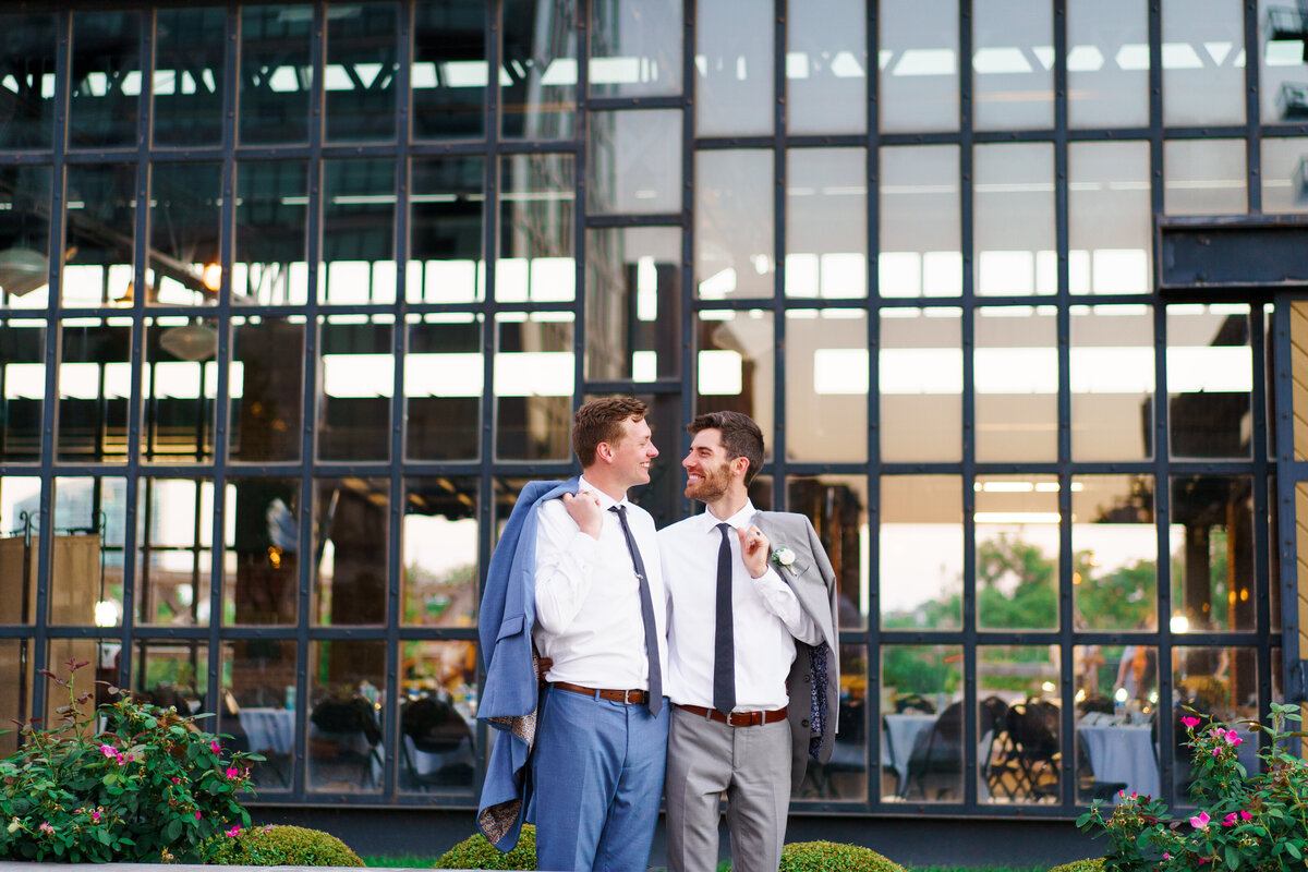 Two grooms smile at each other as they hold their suit jackets off their shoulders in front of a glass wall on their wedding day at North Bank Park Pavilion.