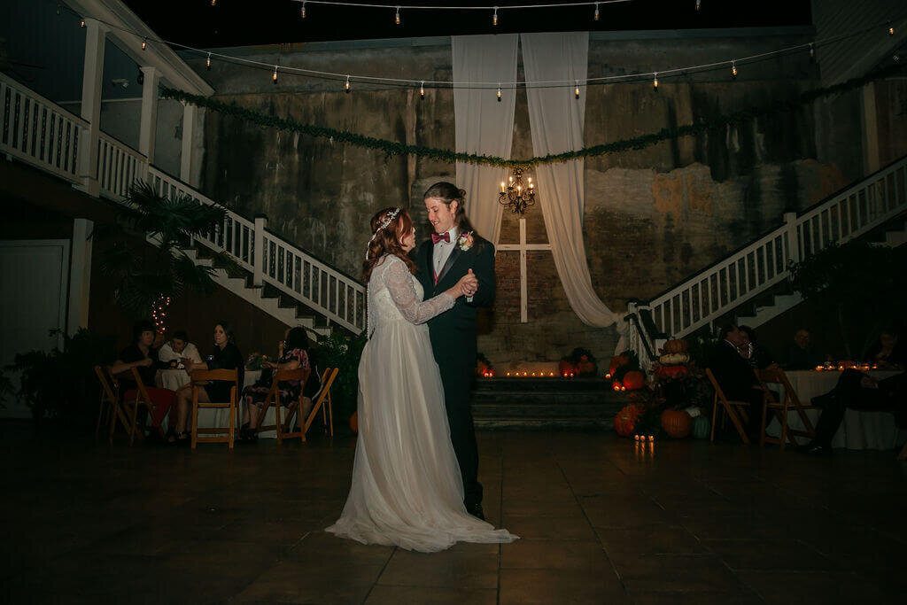 A bride and groom share their first dance in a warmly lit venue with draped fabric, greenery, and pumpkins decorating the space. Guests are seated at tables in the background, watching the couple.