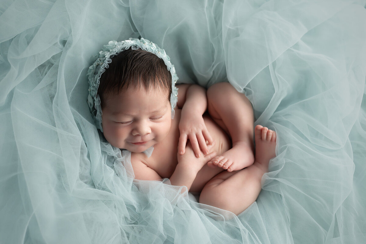 Smiling newborn baby girl in blue floral bonnet, wrapped in blue tulle fabric during a Dallas newborn photo session