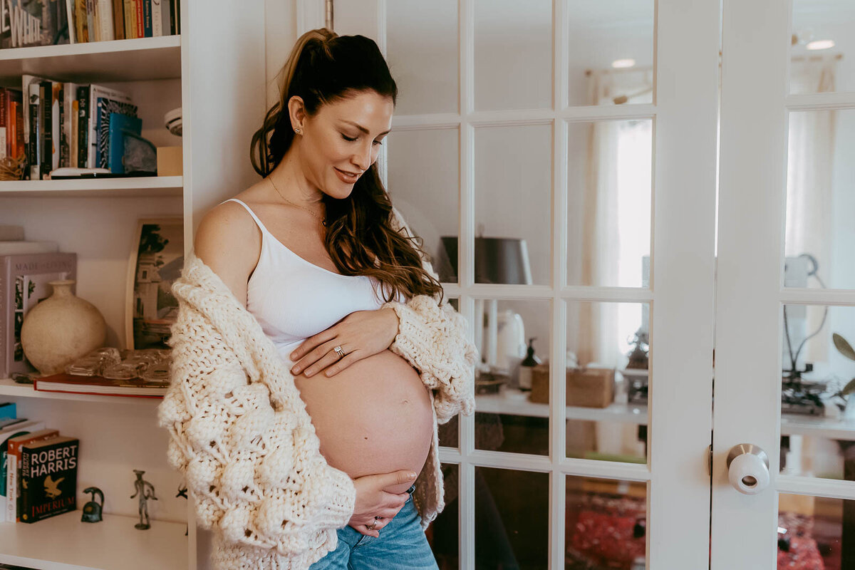 Pregnant woman in a cozy sweater gently cradles her belly, standing near a bookshelf and glass-paned door, smiling softly.