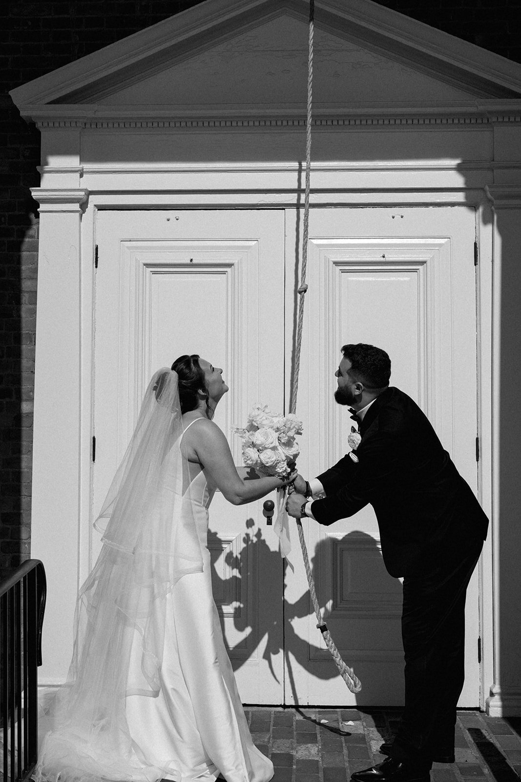 Newlyweds ringing the historic bell outside Martha-Mary Chapel in Dearborn Village Michigan after their wedding ceremony, joyful candid moment.