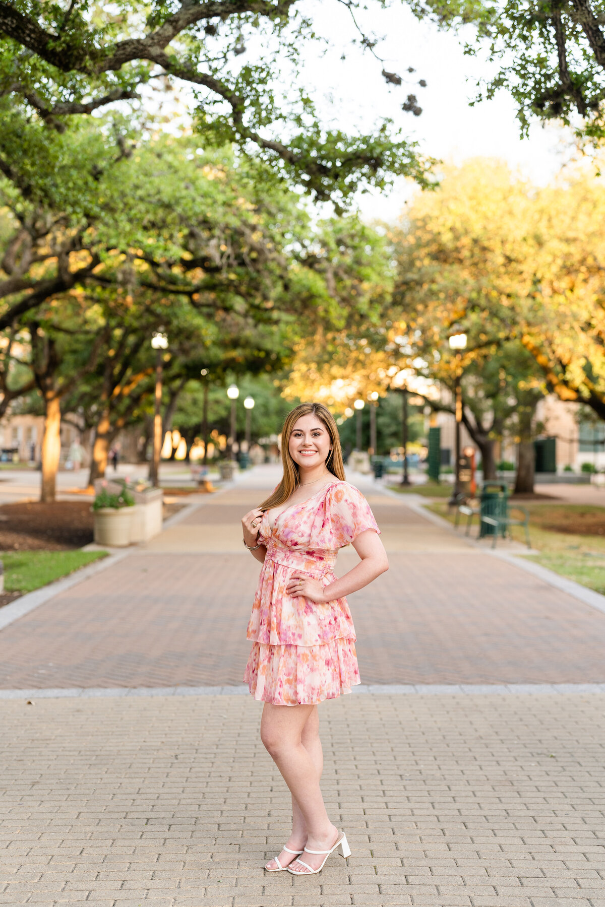 Texas A&M senior girl standing with hand on hip and touching hair while wearing flowery dress in Military Plaza