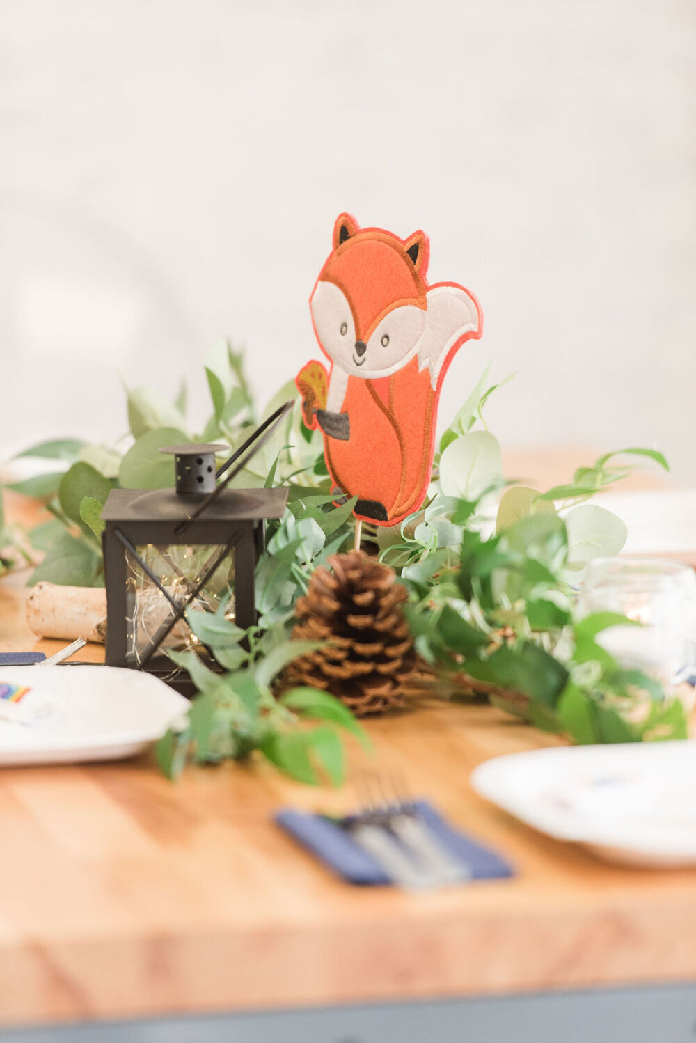 Woodland-themed table centerpiece featuring a charming squirrel perched on wooden logs, surrounded by greenery, pinecones, and a rustic lantern, designed for a baby boy shower.