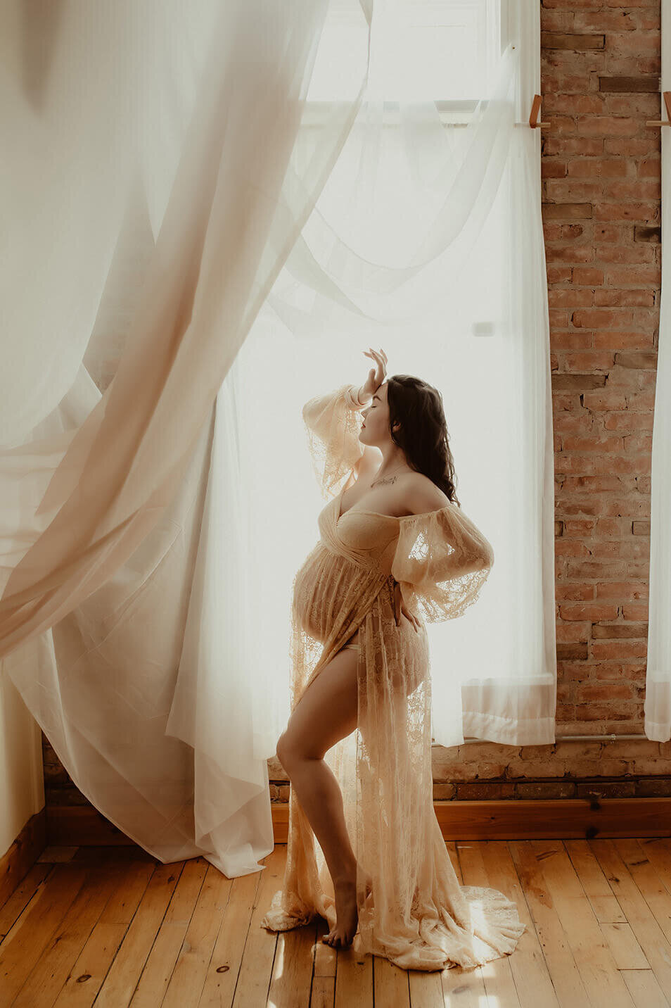 A stunning expectant  mother wearing a cream lace dress standing in front of a window with her hand on her forehead