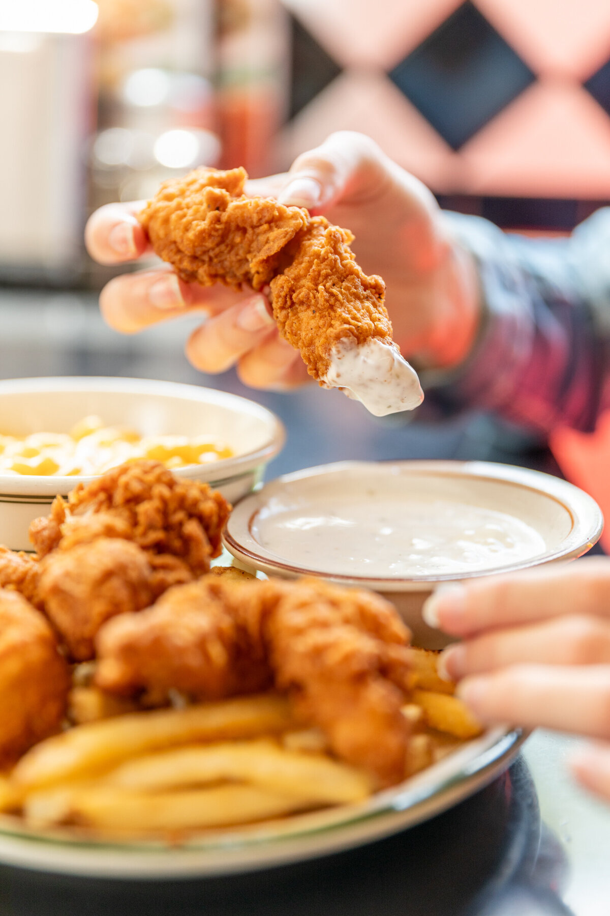 A chicken strip being dipped in a side of Ranch Dressing on a plate with french fries and a side of mac n' cheese.