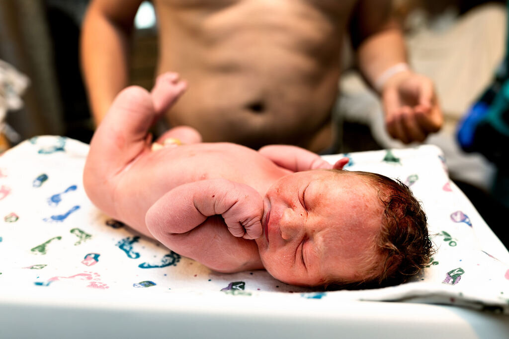 A baby lies on the warmer after being born, his father standing in the background, watching as the doctors prepare to examine him