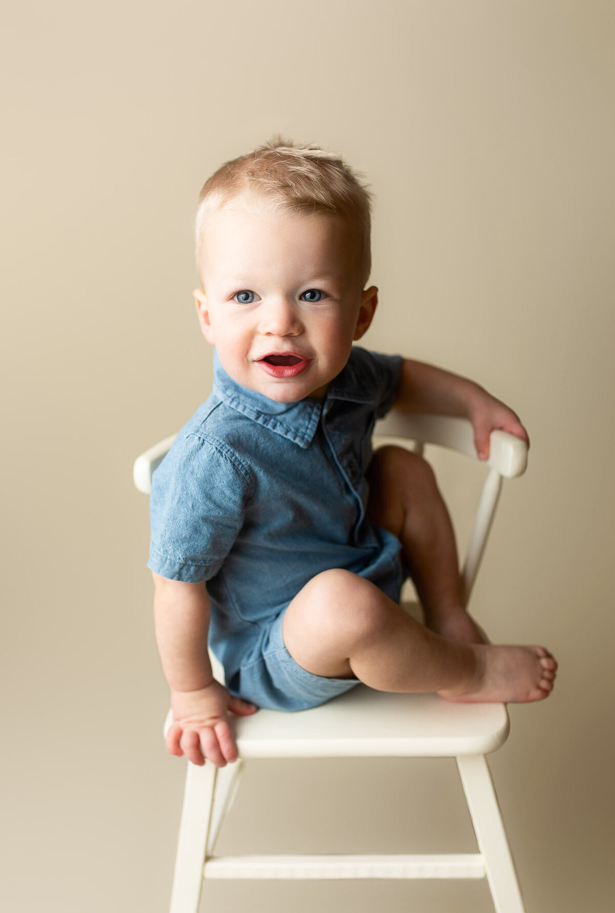 Little blonde boy wearing all denim sitting on a little cream chair with his feet up, cream backdrop in studio