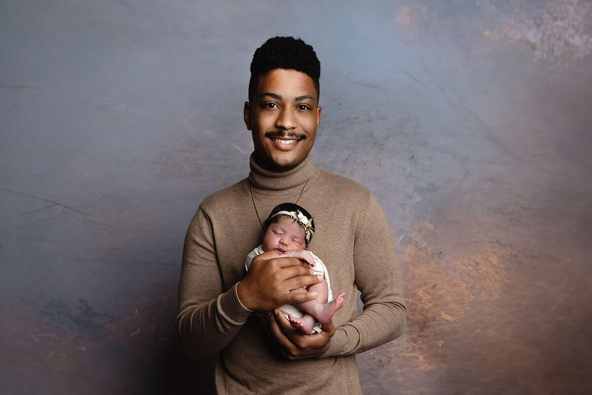 A father in a tan shirt stands smiling in a studio holding his newborn baby