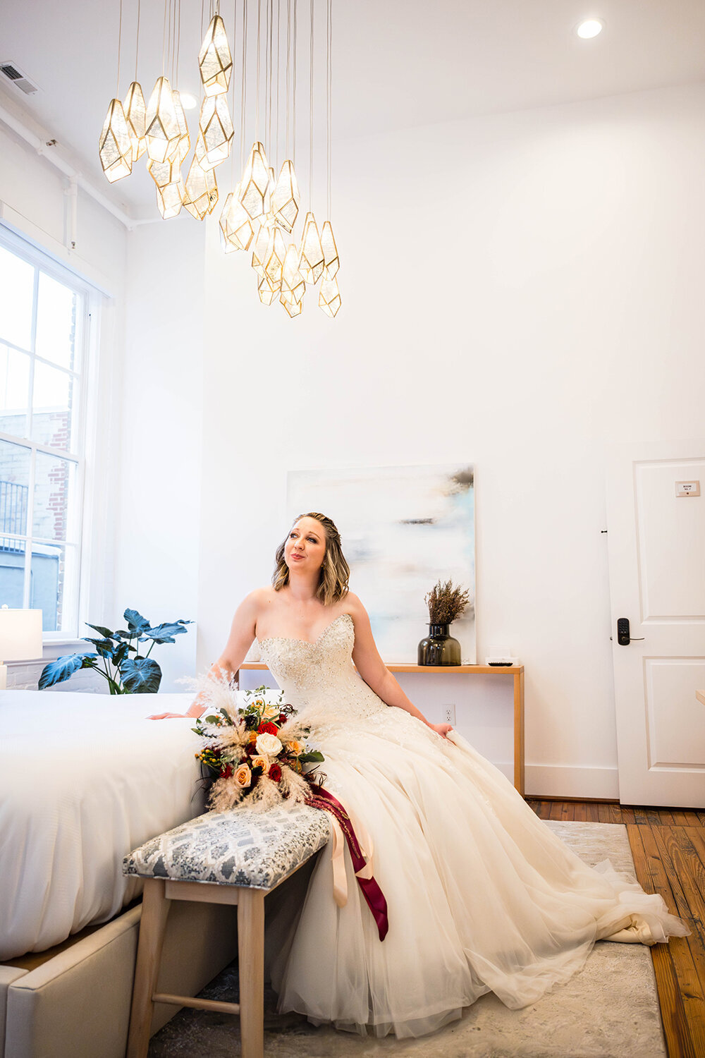 A bride sits on a bench with her wedding bouquet at the foot of a bed in a hotel room at Fire Station One in Roanoke, Virginia for a formal portrait.