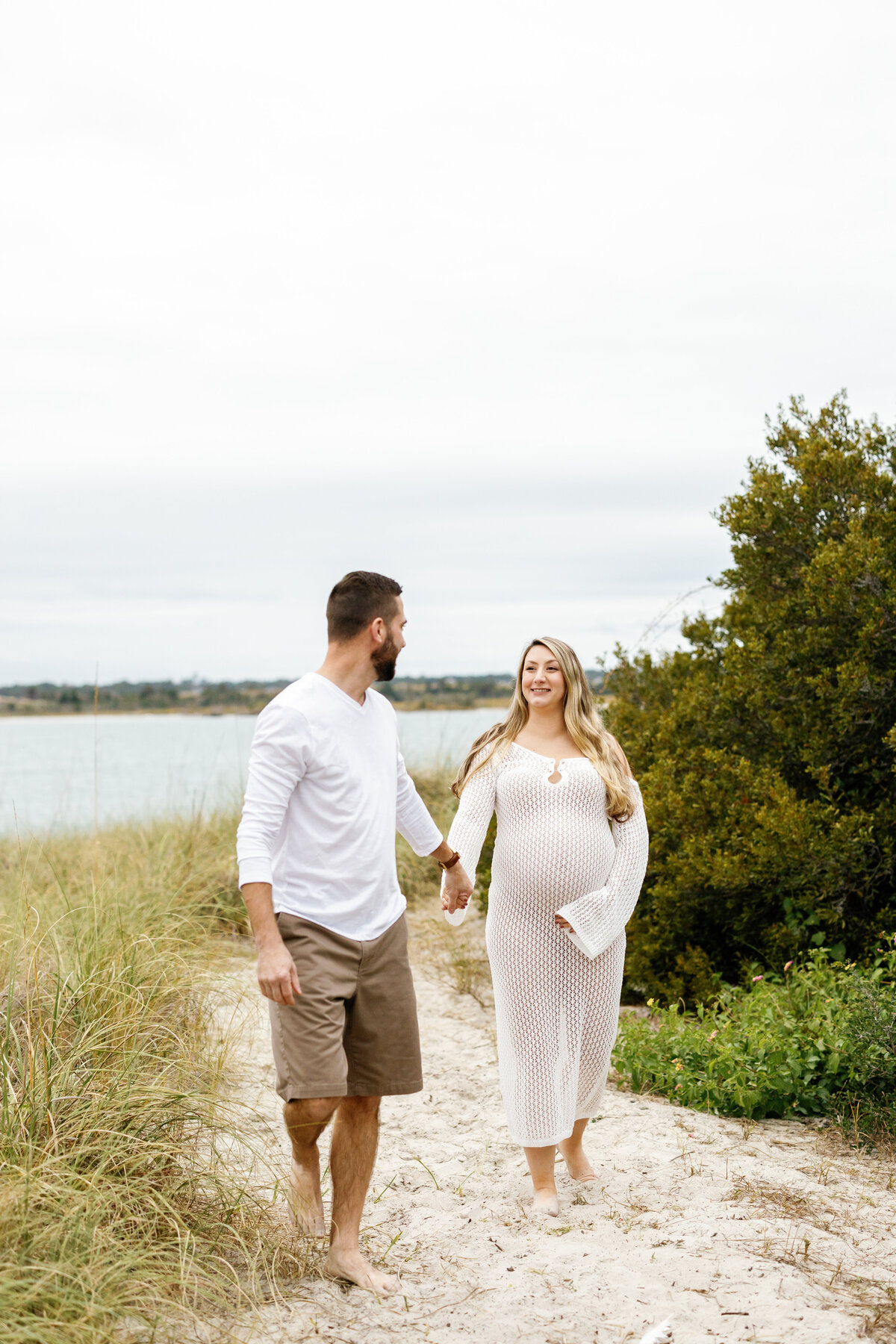 Expectant couple walking hand in hand along Wilmington shoreline, woman's baby bump visible