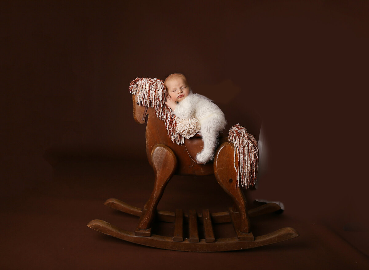 Baby boy is laying atop of a vintage wood rocking horse for his brooklyn newborn photoshoot. one of baby's legs is draping over the side of the horse. Baby's hands are resting atop of the horses mane and baby is sleeping.