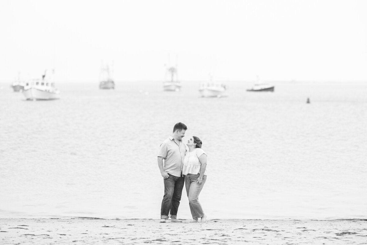 Black-and-white image of a couple standing barefoot on the beach, smiling at each other with fishing boats floating in the background near Chatham Bars Inn in Chatham, Massachusetts — representing the portfolio of a Cape Cod engagement photographer.