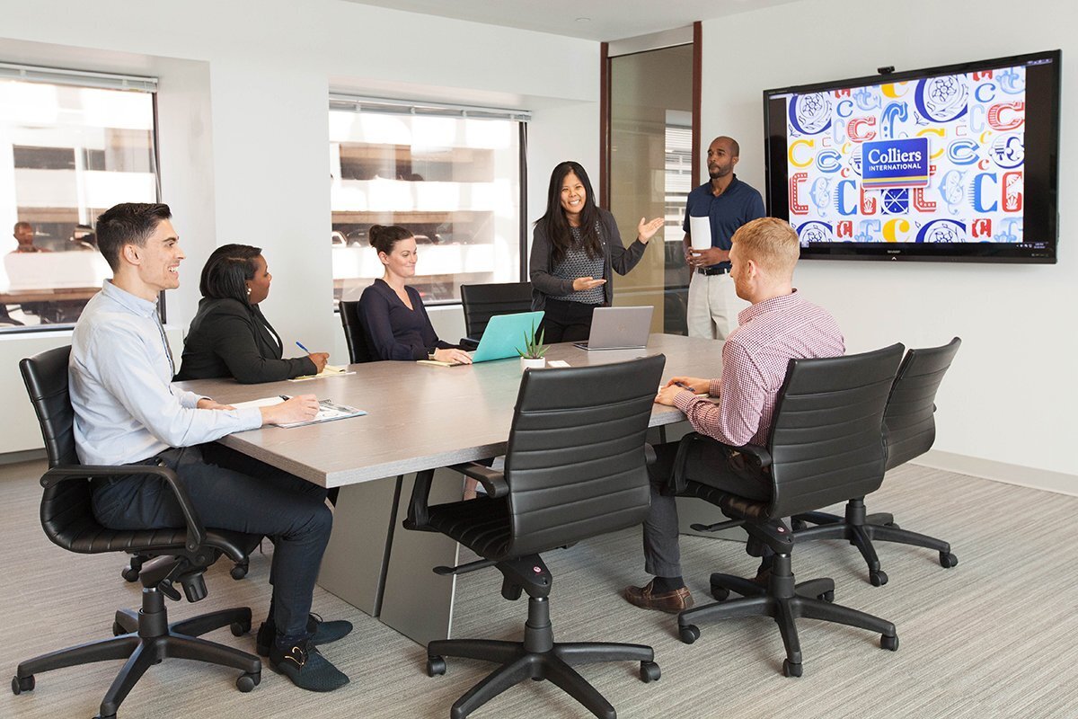 Group of professionals sitting around a boardroom table with woman speaking to them and gesturing towards a large screen with the company logo on it