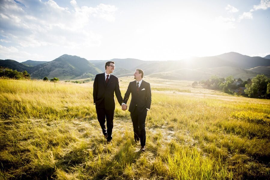 Two grooms walk hand-in-hand toward the camera in a golden field at The Manor House.