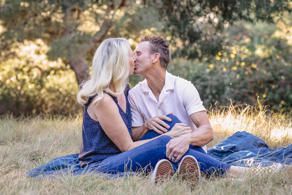A woman with platinum blonde hair, kissing her husband on a navy picnic blanket in green grass, surrounded by greenery at a park in Irvine.
