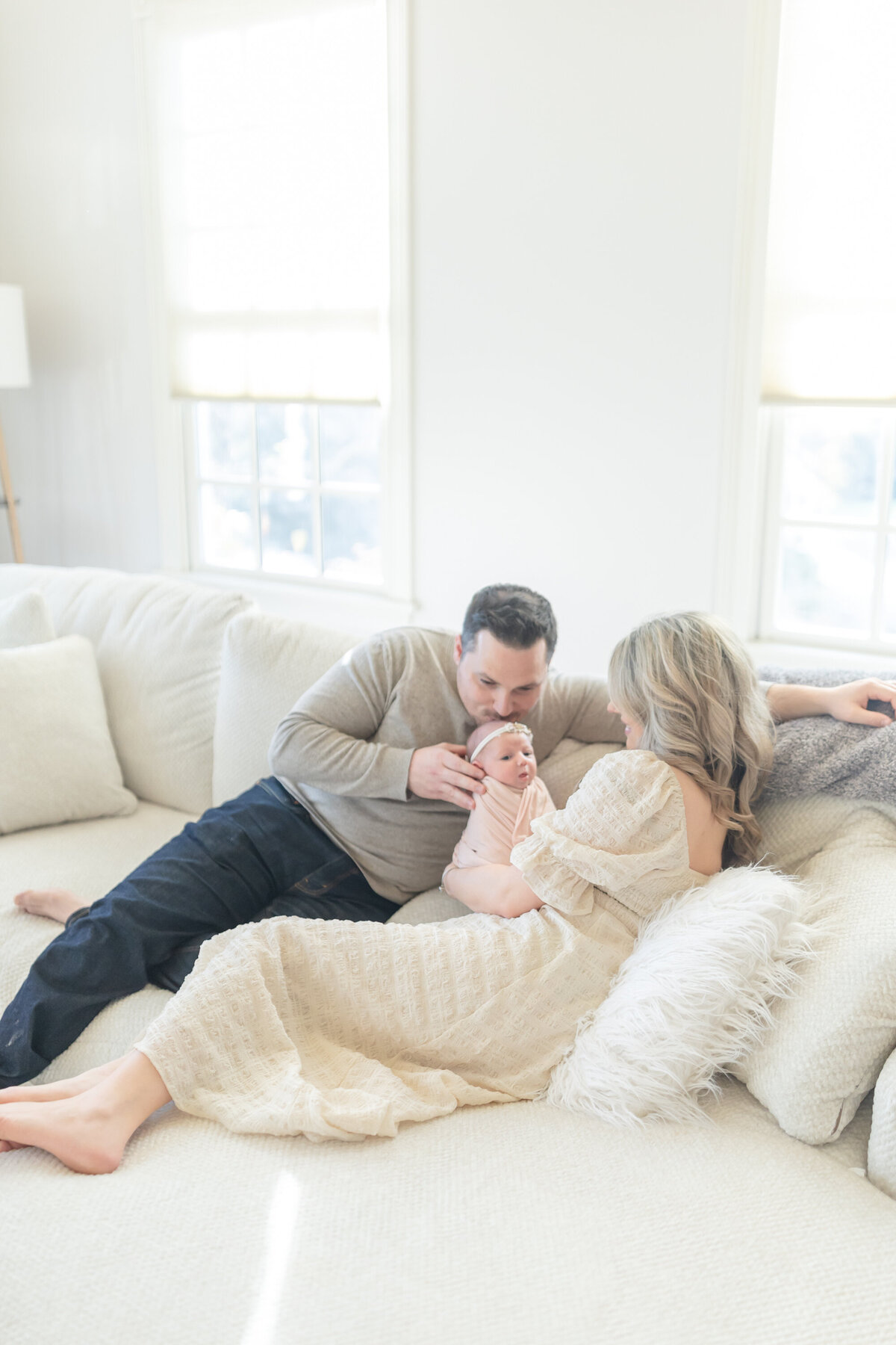 Mom and dad sitting on a cozy couch, lovingly snuggling their newborn baby in an intimate in-home lifestyle session. The parents share a peaceful, tender moment with their little one, surrounded by soft, neutral tones in the room