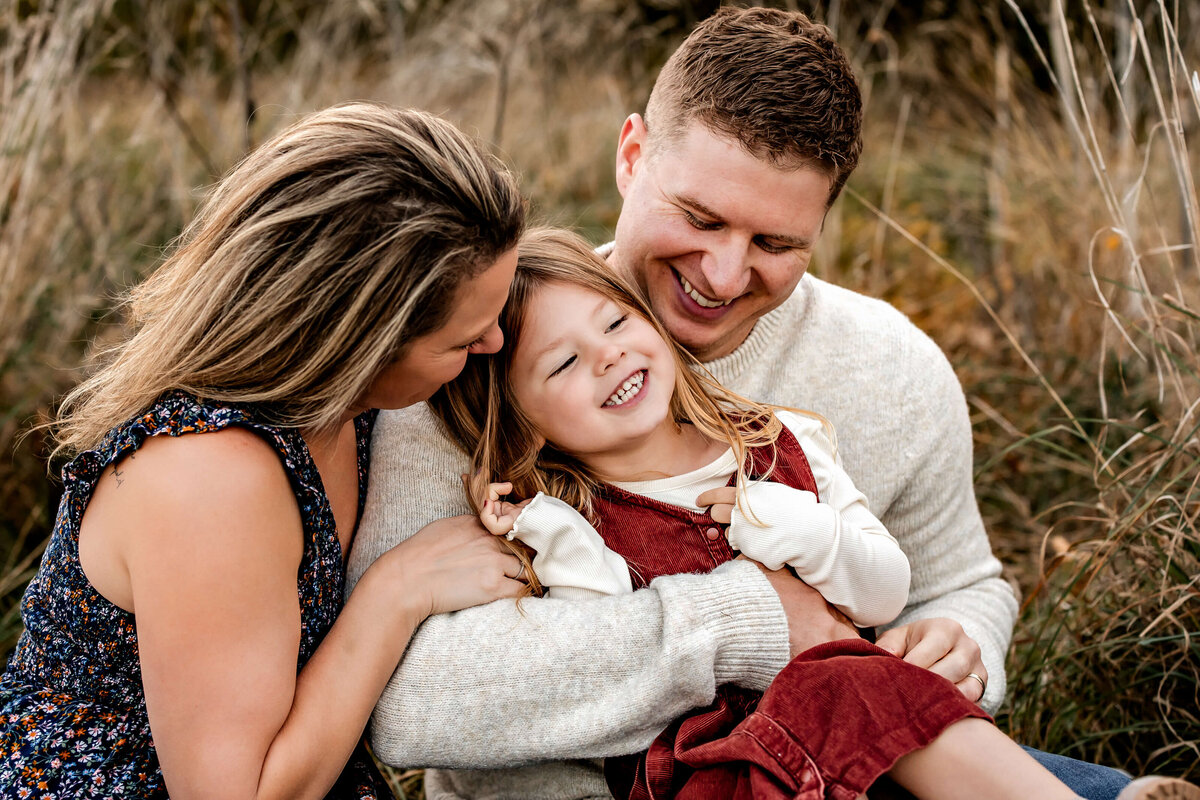 A smiling family sits together outdoors. A man and woman are holding a joyful young girl in their arms. The woman is wearing a patterned dress, the man a light sweater, and the girl a white shirt with a red dress. They are surrounded by tall grass.