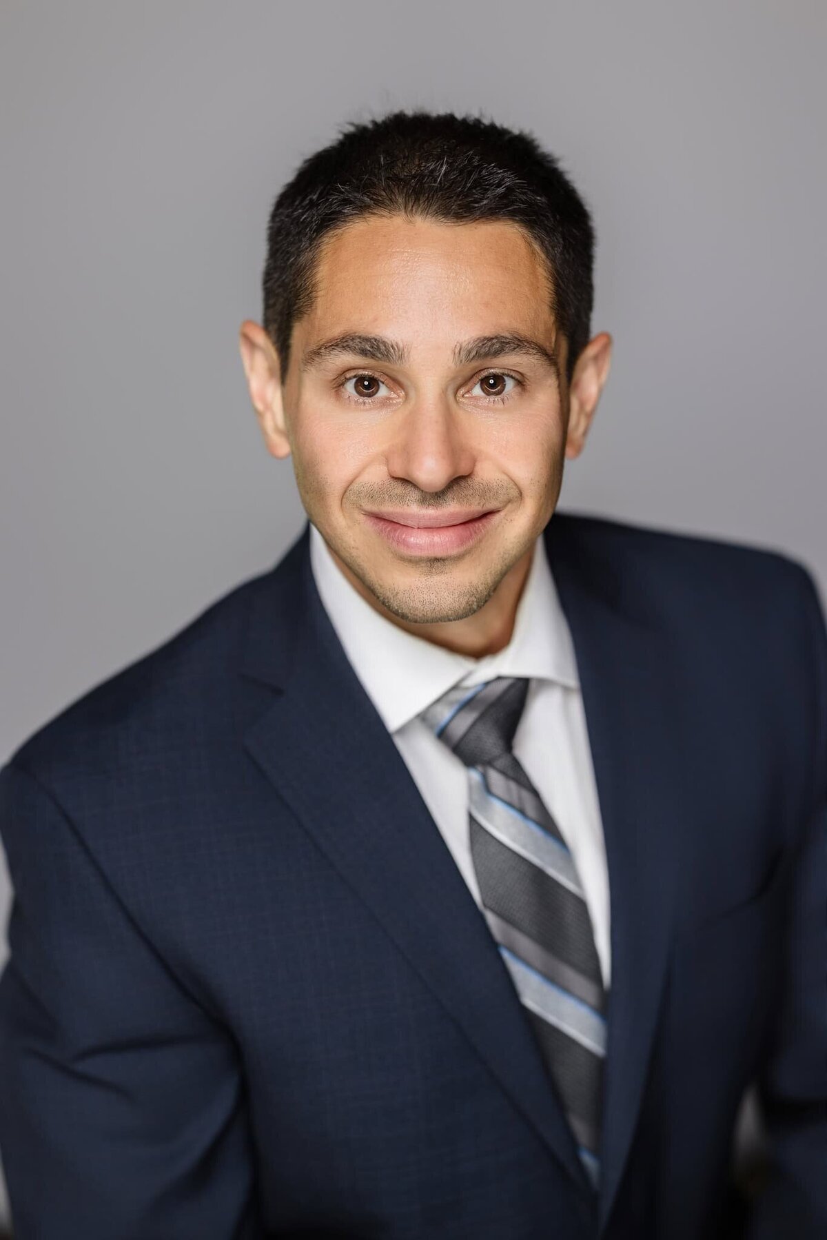 Professional headshot of a man in a navy suit and striped tie smiling on a gray background, Long Island corporate photography.