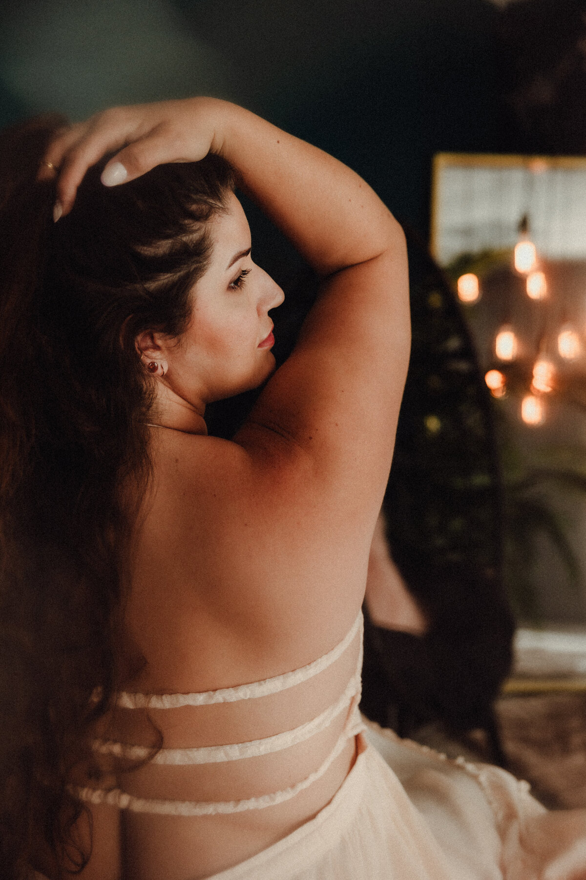 Woman with long dark hair and bold white nails poses with one arm raised, showing her profile and tattooed side in soft studio lighting.