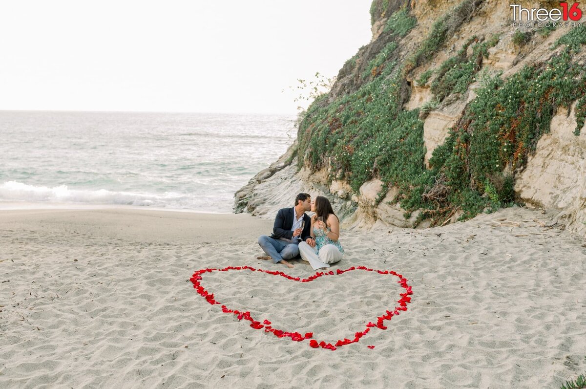 Future Bride and Groom sit on the beach next to roses designed as a heart kissing while holding champagne glasses