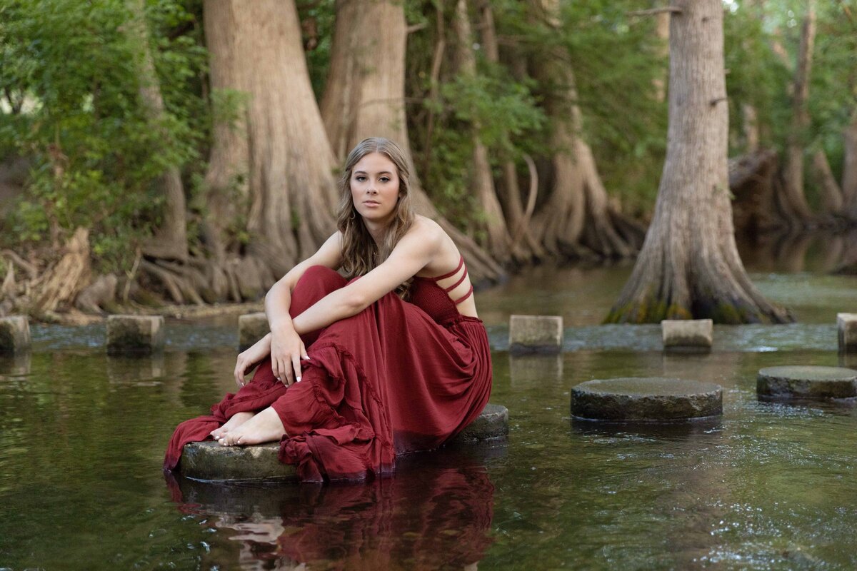 senior girl in a long red dress sitting on a stone slab in the river