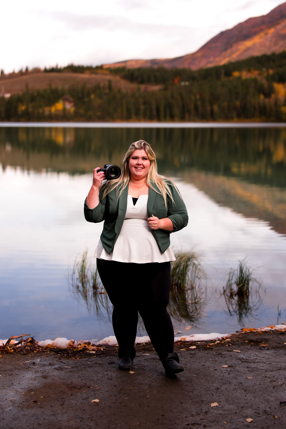 Photographer hold camera in hand and smiles at the camera in front of a lake in the mountains