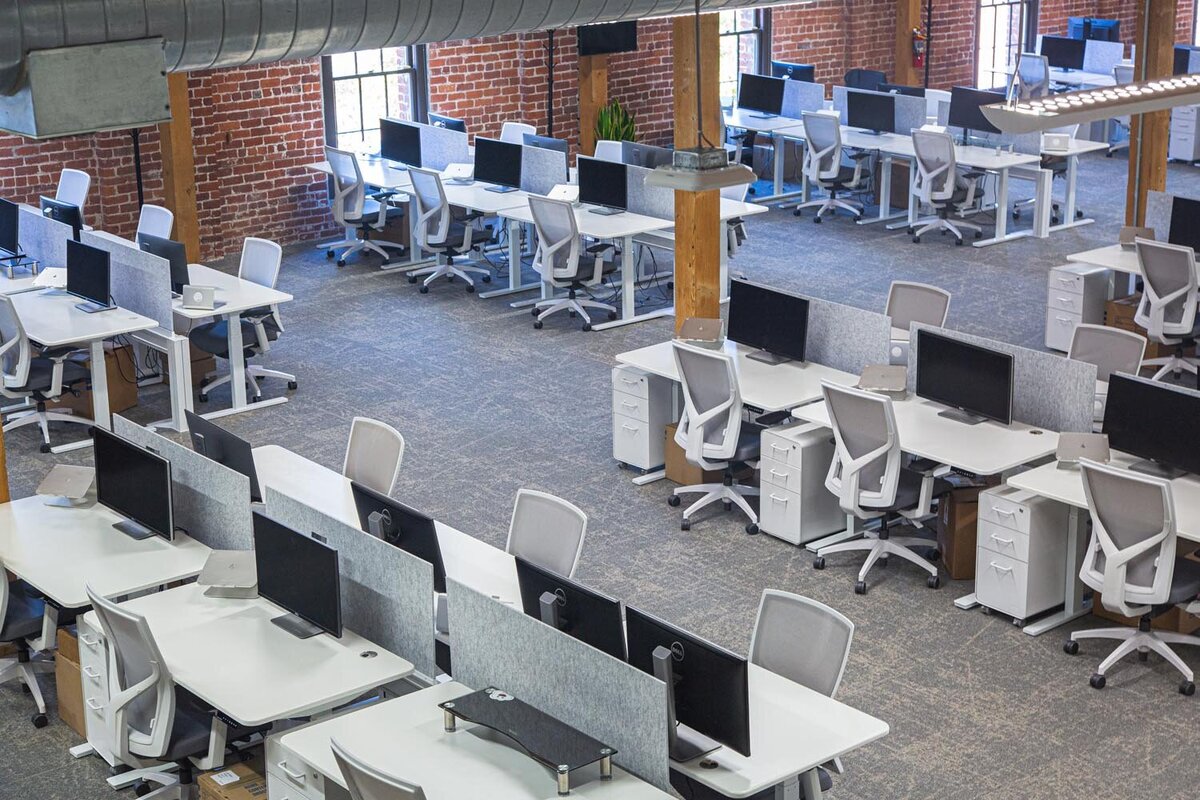 Large open office floor with rows of white desks, ergonomic chairs, and exposed brick walls.