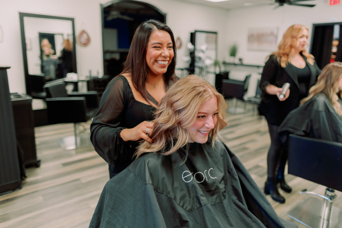 A hairstylist fluffs a client's hair, both are smiling.
