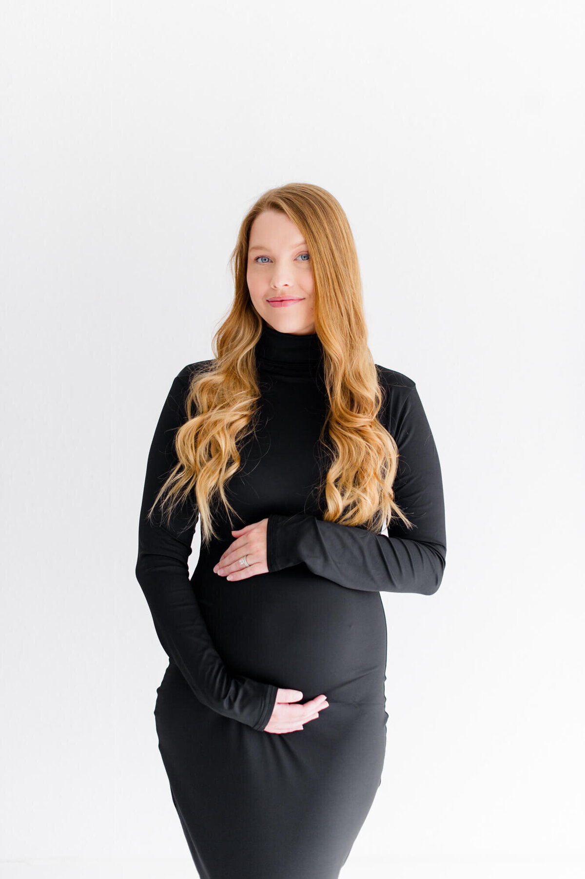 Pregnant mom standing on a white wall holding her belly in a beautiful black dress