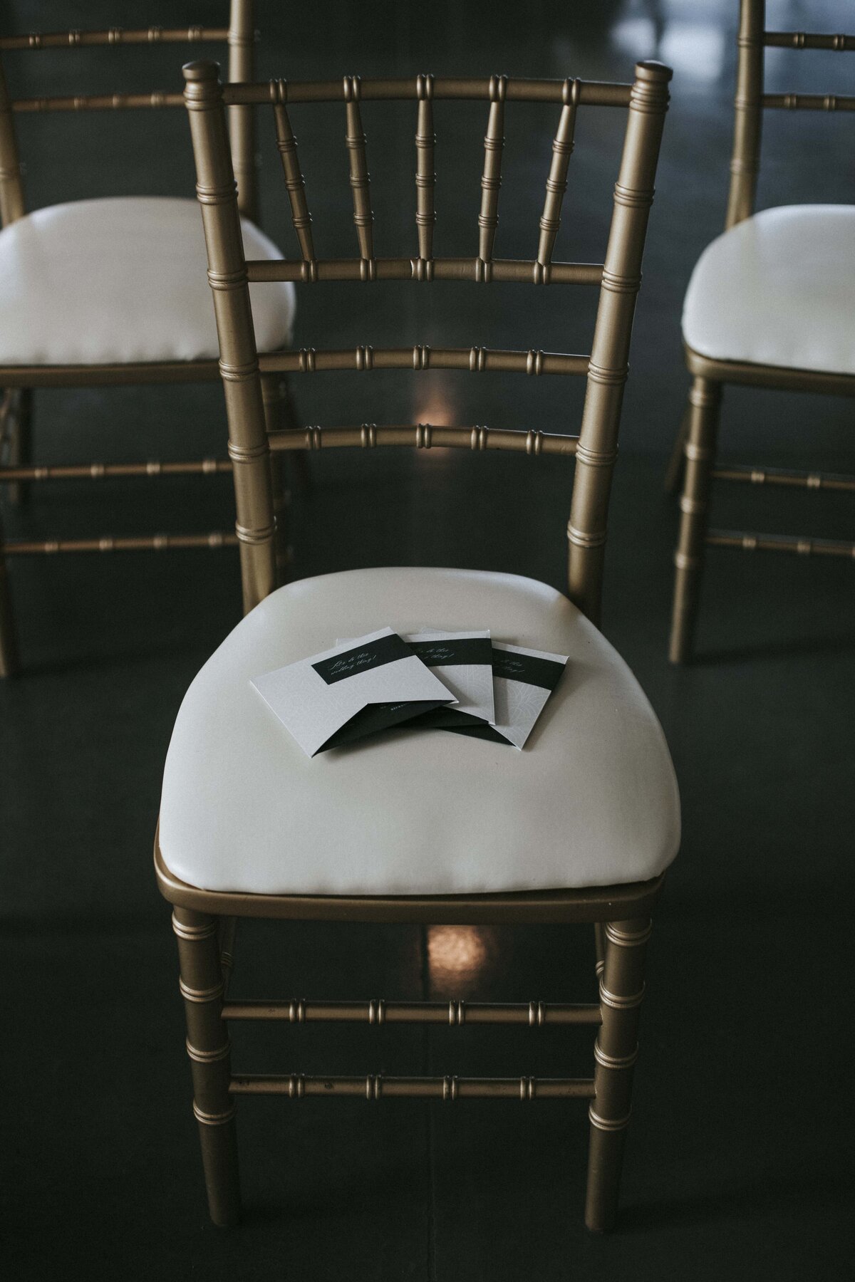 White squared wedding invitations with dark green labels on a gold chair with ivory seat.
