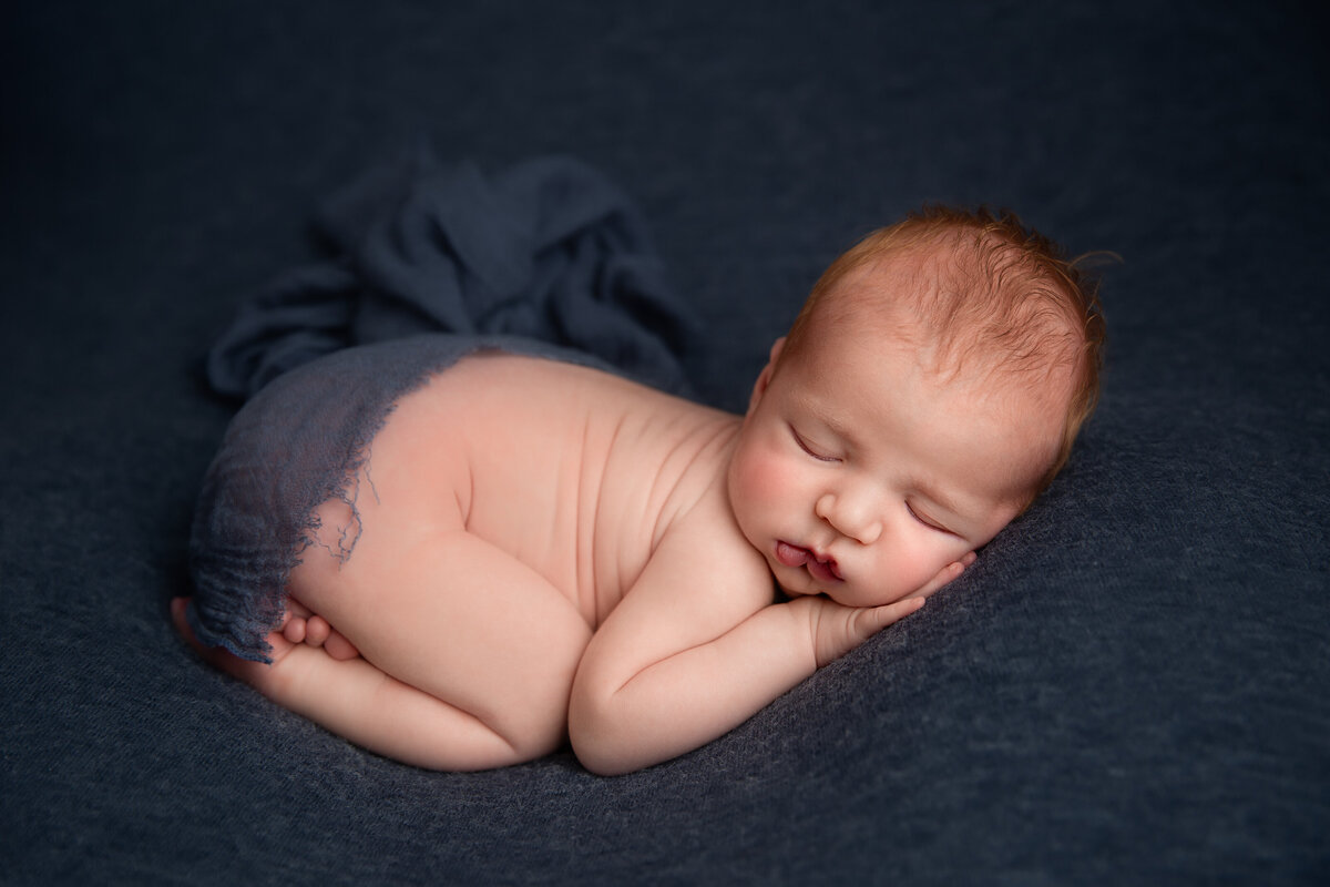 Newborn baby boy sleeping curled up on his stomach on a navy blue fabric background.