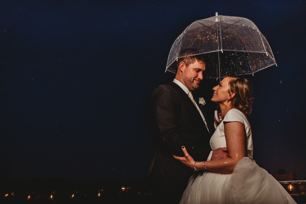 Bride and groom posing for their outdoor wedding under a clear umbrella at night time as the embrace and smile to one another captured by Marilyn wedding photographer