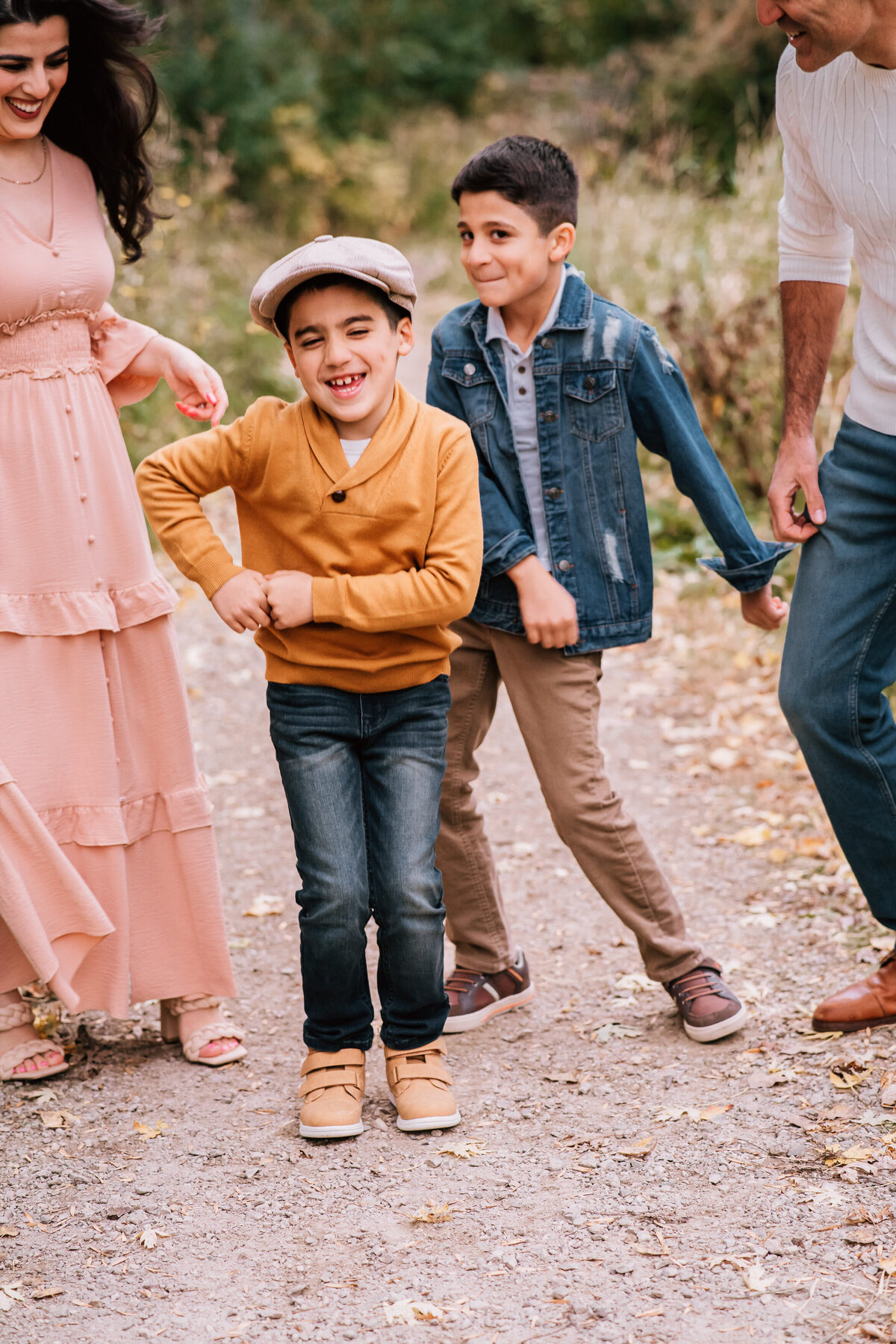 A family moment of joy as a young boy in a yellow sweater and newsboy cap dances playfully, surrounded by his smiling parents and brother on a nature trail.