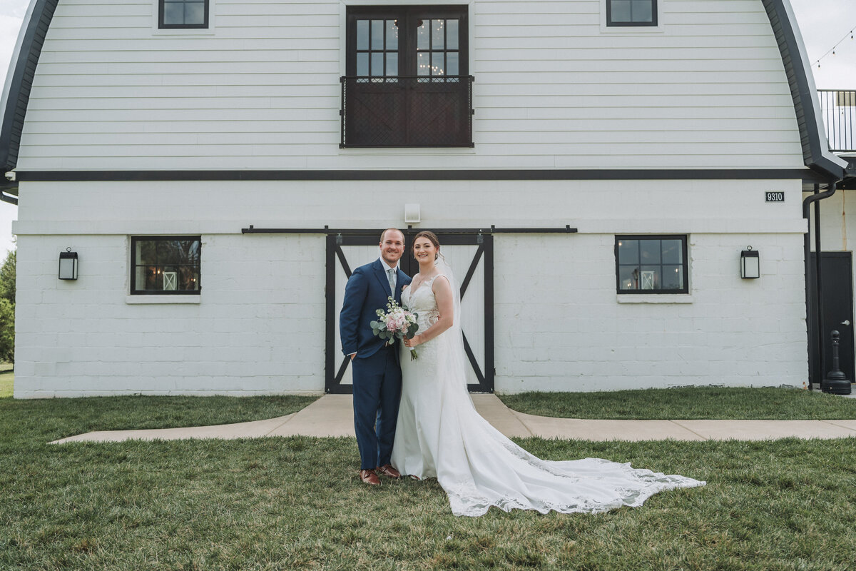 A happy wedding couple stands smiling in front of the iconic white barn at Sweeney Barn in Manassas, Virginia, with the bride holding a floral bouquet, captured by Quinn Photography LLC.